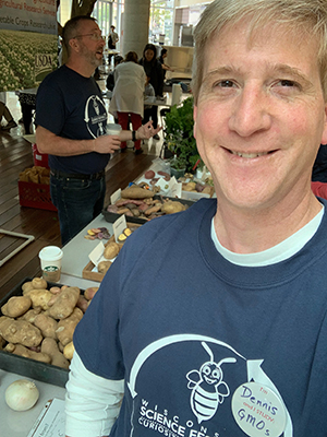 Smiling man standing in front of a table of potatoes, with people talking in the background. He is wearing a button.